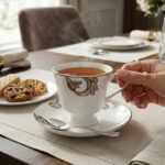A luxury white super bone china cup and saucer featuring gold leaf designs and a gold rim, set on a white placemat on a wooden dining table.