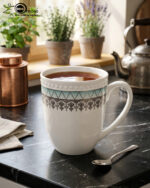 A premium bone china mug filled with hot tea on a black marble countertop, featuring a turquoise geometric pattern and silver lace design, set against a cozy kitchen background with soft natural lighting
