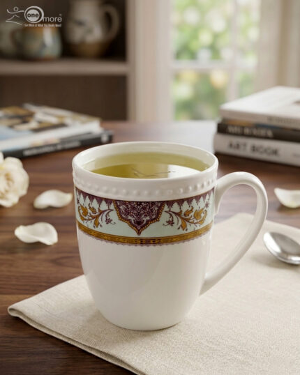 A high-angle close-up of a white bone china mug filled with green tea, featuring an intricate burgundy and gold damask pattern on a mint-green band, resting on a beige linen napkin on a wooden table with books and white rose petals in the background