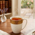 A set of elegant white bone china mugs with an embossed diamond pattern and a golden basket-weave rim, filled with steaming hot tea on a wooden table.