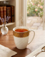 A set of elegant white bone china mugs with an embossed diamond pattern and a golden basket-weave rim, filled with steaming hot tea on a wooden table.