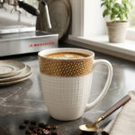 A high-quality bone china coffee mug with a latte inside, featuring a gold woven pattern at the top and an embossed white grid texture on the bottom, set on a dark marble countertop next to coffee beans and a gold spoon