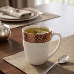 A luxury white bone china mug with a rose gold mesh design on the rim, filled with steaming green tea and a tea leaf floating on top, placed on a textured placemat with a silver spoon on a wooden dining table