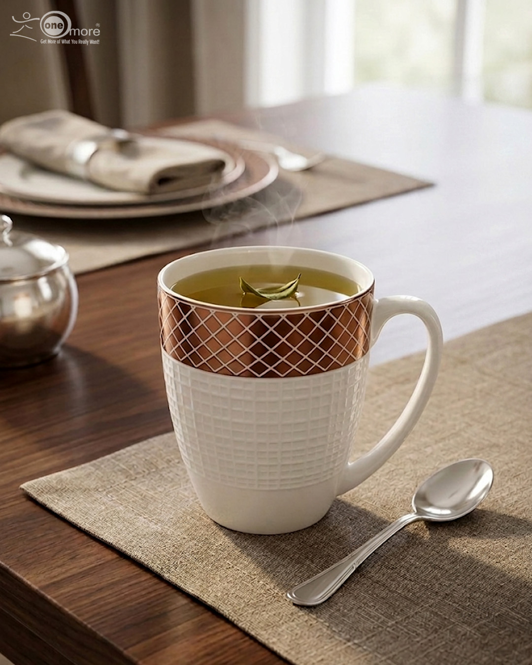 3 A luxury white bone china mug with a rose gold mesh design on the rim, filled with steaming green tea and a tea leaf floating on top, placed on a textured placemat with a silver spoon on a wooden dining table
