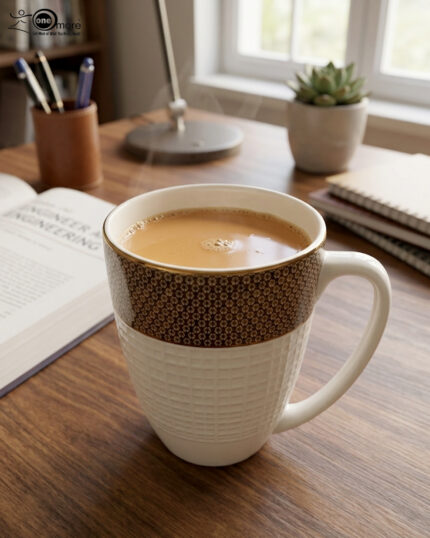 A high-angle close-up of a premium bone tea mug filled with hot tea, featuring a gold-rimmed floral pattern and a white textured base on a wooden desk with a book and succulent in the background