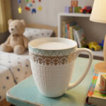 A high-angle close-up of a white bone china mug filled with milk, featuring a gold filigree border and textured lattice design, resting on a rustic blue wooden table in a brightly lit room