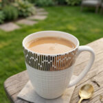 A high-angle shot of a white bone china mug filled with tea, featuring a gold geometric pattern on a wooden outdoor table with a gold spoon and green garden background