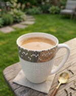 A high-angle shot of a white bone china mug filled with tea, featuring a gold geometric pattern on a wooden outdoor table with a gold spoon and green garden background