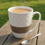 A set of six premium white ceramic tea mugs with a textured lattice design and a brown patterned base, filled with steaming tea and placed on a wooden outdoor table next to a silver spoon