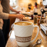 A close-up shot of a white bone china mug with an embossed diamond pattern and a brown Greek key design, filled with latte art, sitting on a granite kitchen countertop next to a silver spoon