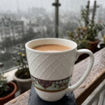 A close-up shot of a white bone china mug filled with tea, resting on a slate coaster on a rainy balcony railing. The mug features an embossed diamond pattern and a decorative floral band at the bottom