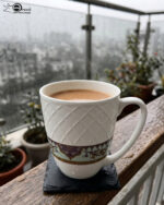 A close-up shot of a white bone china mug filled with tea, resting on a slate coaster on a rainy balcony railing. The mug features an embossed diamond pattern and a decorative floral band at the bottom