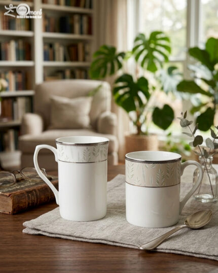 A professional lifestyle shot (3.jpg) showcasing two white Bone China mugs with silver rims and a green botanical leaf pattern, featuring one tall "long" mug and one "short" mug on a linen cloth in a cozy living room setting