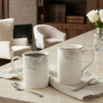 A high-angle shot labeled 3.jpg showing two elegant white bone china mugs with floral patterns and silver rims on a marble countertop next to silver spoons