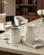 A high-angle shot labeled 3.jpg showing two elegant white bone china mugs with floral patterns and silver rims on a marble countertop next to silver spoons