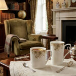 A professional product shot labeled 3.jpg showing two elegant bone china mugs with floral patterns and gold rims on a wooden table, set against a classic living room background with an armchair and fireplace