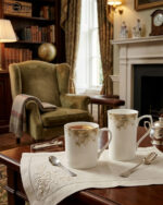 A professional product shot labeled 3.jpg showing two elegant bone china mugs with floral patterns and gold rims on a wooden table, set against a classic living room background with an armchair and fireplace