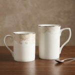 A set of two white bone china mugs on a wooden surface, one short and one tall, featuring elegant gold lace patterns and metallic gold rims against a neutral background.