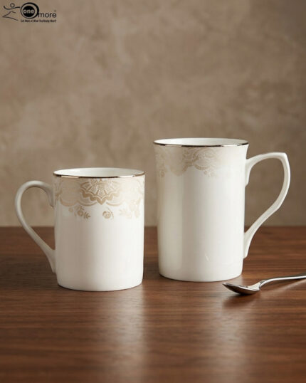 A set of two white bone china mugs on a wooden surface, one short and one tall, featuring elegant gold lace patterns and metallic gold rims against a neutral background.