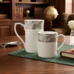 Set of two white bone china mugs from 3.jpg on a wooden table with a green placemat; one tall mug and one short mug, both featuring a grey marble pattern band and silver trim against a library background with cinematic lighting