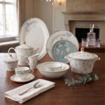 A luxury 61-piece Super Bone China dinner set with blue floral patterns and silver rims displayed on a wooden dining table, including plates, bowls, a teapot, and a serving casserole.