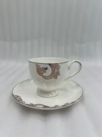 A steaming stream of milk tea being poured into a luxury white Super Bone China cup with gold-leaf designs, resting on a matching saucer with a silver spoon on a dark wooden table