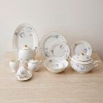 A complete super bone china dinner set in file 3.jpg featuring a delicate blue rose pattern, gold-rimmed edges, and including a teapot, plates, bowls, and serving dishes on a marble surface.