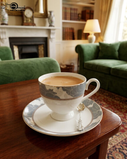 A high-angle shot of a premium super bone china cup and saucer filled with milk tea, featuring a grey floral pattern and silver spoon, set on a wooden table in a cozy living room with green sofas and a fireplace.