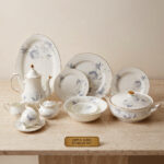 An elegant, white super bone dinner set with a delicate blue rose floral pattern and gold rims, displayed on a beige marble tabletop against a neutral background. The set includes dinner plates, side plates, a large oval serving platter, a covered casserole dish, a teapot, a sugar bowl, a creamer pitcher, a teacup with a saucer, and serving bowls