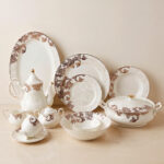 An elegant white super bone china dinner set featuring a classic brown and gold baroque acanthus leaf pattern, including plates, bowls, a teapot, a tureen, and teacups arranged on a light-colored surface