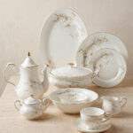 An elegant white super bone china dinner set featuring delicate beige floral patterns and gold rims, arranged on a textured beige surface. The set includes plates, bowls, a teapot, a covered casserole dish, a cream pitcher, a sugar bowl, and a teacup with a saucer.