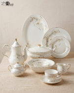 An elegant white super bone china dinner set featuring delicate beige floral patterns and gold rims, arranged on a textured beige surface. The set includes plates, bowls, a teapot, a covered casserole dish, a cream pitcher, a sugar bowl, and a teacup with a saucer.
