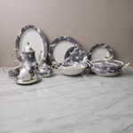 A luxury grey and white floral pattern bone china tea set and plates arranged on a wooden table, including a teapot, teacup with saucer, creamer, sugar bowl, and dinner plates.