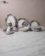 A luxury grey and white floral pattern bone china tea set and plates arranged on a wooden table, including a teapot, teacup with saucer, creamer, sugar bowl, and dinner plates.