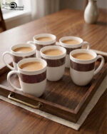A set of six elegant white bone china mugs with a decorative rose-gold geometric band and beaded rim, displayed on a wooden tray with hot tea and rising steam in a warm, cinematic library setting