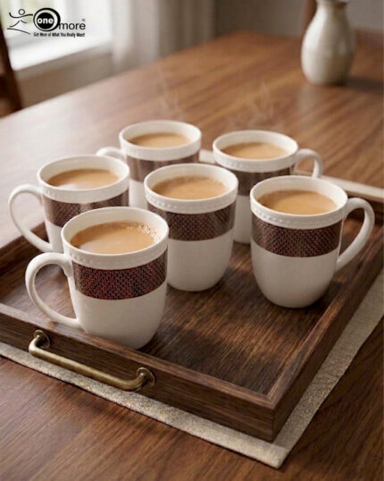 A set of six elegant white bone china mugs with a decorative rose-gold geometric band and beaded rim, displayed on a wooden tray with hot tea and rising steam in a warm, cinematic library setting