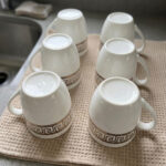 A high-quality white bone china mug filled with steaming milk tea, featuring a gold Greek Key pattern and beaded texture, resting on a rustic wooden garden table