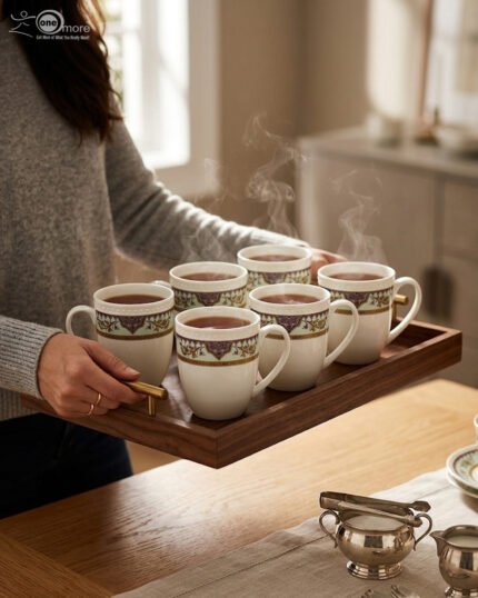 A high-angle close-up of a white bone china mug filled with green tea, featuring an intricate burgundy and gold damask pattern on a mint-green band, resting on a beige linen napkin on a wooden table with books and white rose petals in the background