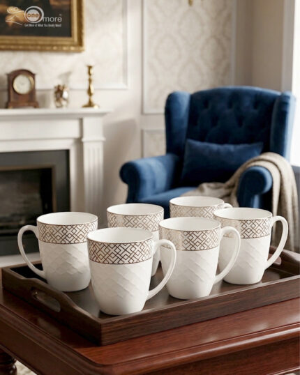 A steaming cup of latte with latte art inside a white bone china mug featuring a gold geometric pattern and quilted texture, resting on a glass saucer with a silver spoon on a wooden table
