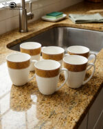 A high-quality bone china coffee mug with a latte inside, featuring a gold woven pattern at the top and an embossed white grid texture on the bottom, set on a dark marble countertop next to coffee beans and a gold spoon