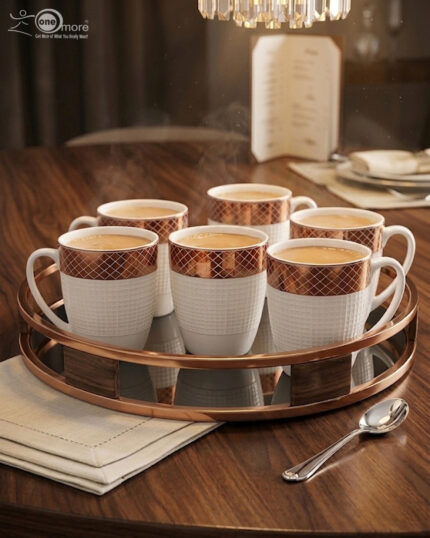 A luxury white bone china mug with a rose gold mesh design on the rim, filled with steaming green tea and a tea leaf floating on top, placed on a textured placemat with a silver spoon on a wooden dining table