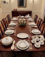 An elegant, white super bone dinner set with a delicate blue rose floral pattern and gold rims, displayed on a beige marble tabletop against a neutral background. The set includes dinner plates, side plates, a large oval serving platter, a covered casserole dish, a teapot, a sugar bowl, a creamer pitcher, a teacup with a saucer, and serving bowls