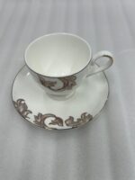 A steaming stream of milk tea being poured into a luxury white Super Bone China cup with gold-leaf designs, resting on a matching saucer with a silver spoon on a dark wooden table