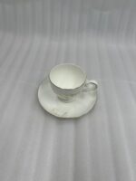 A high-angle shot of a white fine bone china tea cup filled with milk tea, featuring a delicate white floral pattern and gold-trimmed edges, resting on a matching saucer on a wooden side table in a cozy, classic living room setting