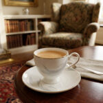 A luxury white super bone china teacup filled with creamy tea, featuring gold-rimmed edges and a delicate grey leaf pattern, resting on a matching saucer on a dark wooden table in a cozy, blurred library background