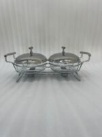 Two-dish Paramount Series stainless steel food warmer with glass bowls and chrome lids, featuring two buffet burners underneath, displayed on a wooden dining table.