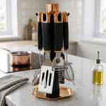A 7-piece kitchen gadget set featuring black matte handles and rose gold accents, displayed on a matching rotating stand atop a white marble kitchen counter.