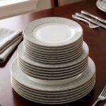 A stacked 18-piece elegant super bone china plate set on a dark wooden dining table. The set includes quarter plates on top, deep plates in the middle, and large rice plates at the bottom, all featuring a light grey floral pattern border and gold trim