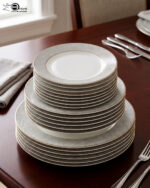 A stacked 18-piece elegant super bone china plate set on a dark wooden dining table. The set includes quarter plates on top, deep plates in the middle, and large rice plates at the bottom, all featuring a light grey floral pattern border and gold trim