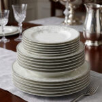A stacked 18-piece elegant white super bone china dinner plate set featuring a delicate grey floral pattern on the rims, neatly arranged on a grey fabric runner over a wooden dining table with crystal glasses and a silver pitcher in the background.